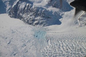 "Aerial view of a glacier flowing between snow-covered mountains into a frozen bay, with chunks of ice and a patch of light blue meltwater visible."