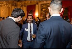 Professionals in formal attire engage in conversation at a high-end Wall Street networking event, set in an opulent, gold-trimmed venue. Despite rising tensions in the Middle East, the mood remains composed, reflecting the financial sector’s current sense of calm amid global uncertainty.